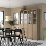 Modern dining area with large wooden BILLY bookcase, black chairs, striped rug. Light wood, glass doors showcase dishes. Simple, clean design.