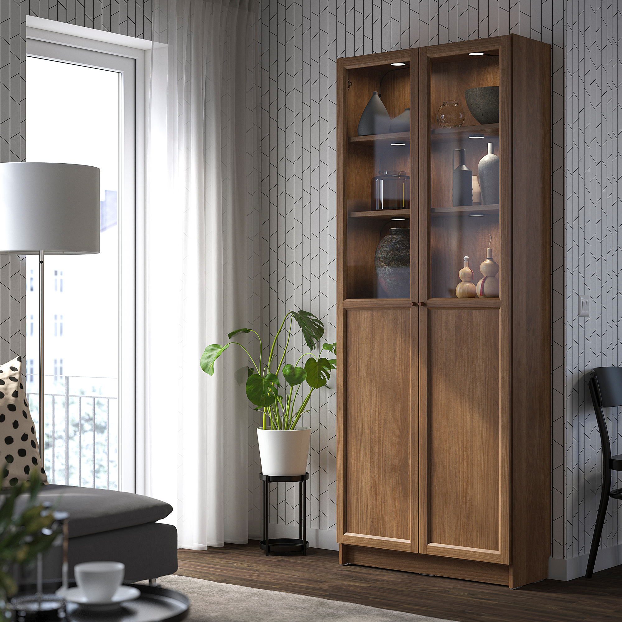 Stylish wooden BILLY bookcase with glass doors in living room, beside a window, holding vases, contrasting gray sofa, warm toned.