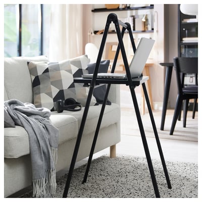 Black adjustable stand-up desk with laptop in living space, next to a gray sofa with a geometric pillow and blanket, offering a comfortable workspace.