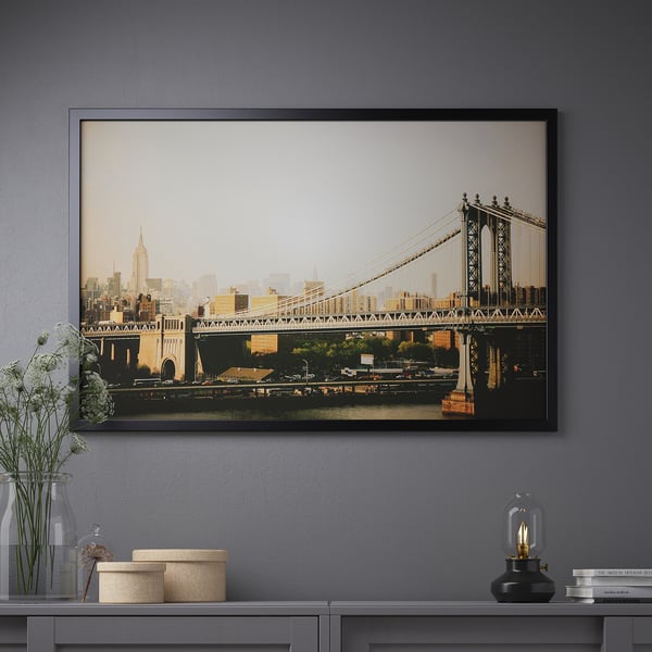 A large framed a cityscape with a bridge. The frame is displayed on a wall above a furniture unit with decorative items.