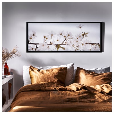 Cozy bedroom with a large brown bedspread and pillows. Above, a framed floral print, BJÖRKSTA, enhances the rooms aesthetic with white flowers.