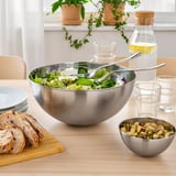 Large stainless-steel bowl with salad, tongs, bread, nuts, glasses, and water jug on a wooden table.