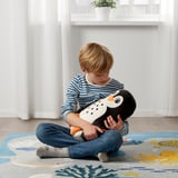 Child hugs penguin cushion on blue rug. Wears striped shirt, jeans. Recycled materials. Plant behind.