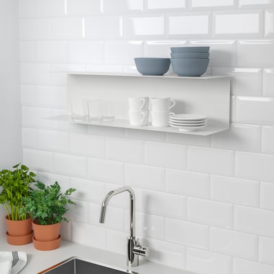Kitchen shelves displaying dishes, glasses, and bowls. White tiled backsplash and chrome faucet. Potted plants by sink.