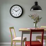 Black BRAVUR wall clock above dining table with flowers and chairs.