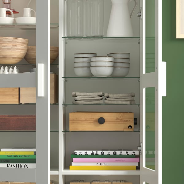 Modern cabinet with glass doors displaying dishes, folded linens, wooden box, and colourful books.