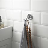 A chrome double hook on a tiled bathroom wall holds a gray towel and a wooden bead string, near a white sink with soap dispenser.
