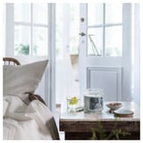 Cozy room with white bed, glass candle, bowl, and lemon drink on table by window.