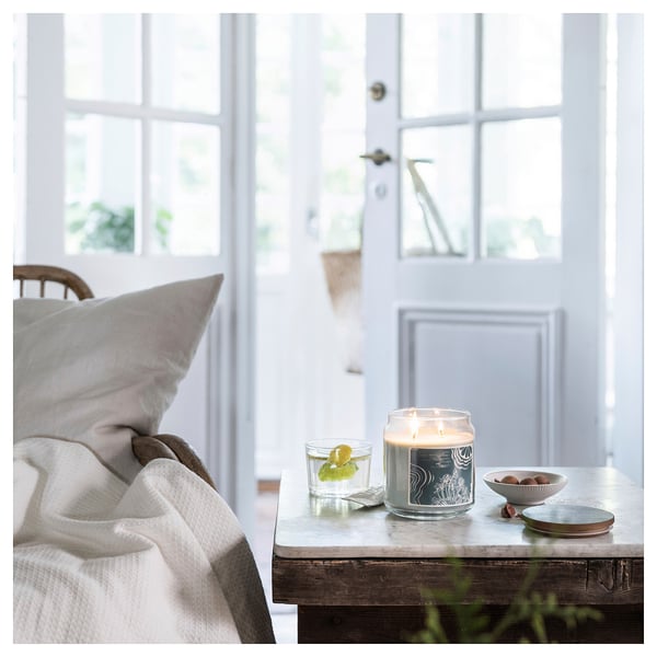 Cozy room with white bed, chair, and nightstand. On nightstand: lit candle, glass with lemon, bowl of nuts, coaster. Door and windows are visible.