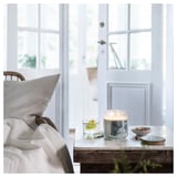 Cozy room with white bed, chair, and nightstand. On nightstand: lit candle, glass with lemon, bowl of nuts, coaster. Door and windows are visible.