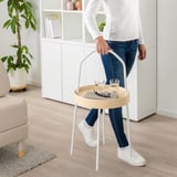 Person holding a round, white side table with a handle, carrying two glasses, a remote, and a book. The table is easy to carry around the room.