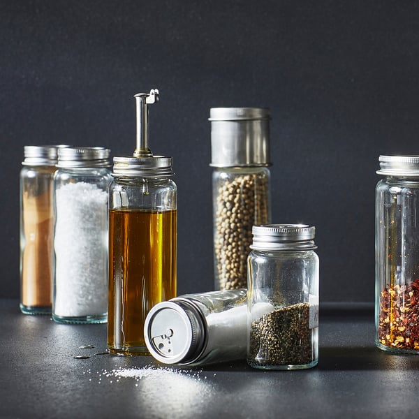 Six glass containers with metal lids and contents including salt, oil, spices, and dried chili flakes, organized neatly on a dark surface.