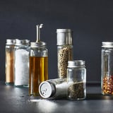 Six glass containers with metal lids and contents including salt, oil, spices, and dried chili flakes, organized neatly on a dark surface.