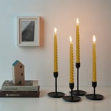 Four lit twisted yellow candles in black holders on a table with a picture frame, books, and a birdhouse.