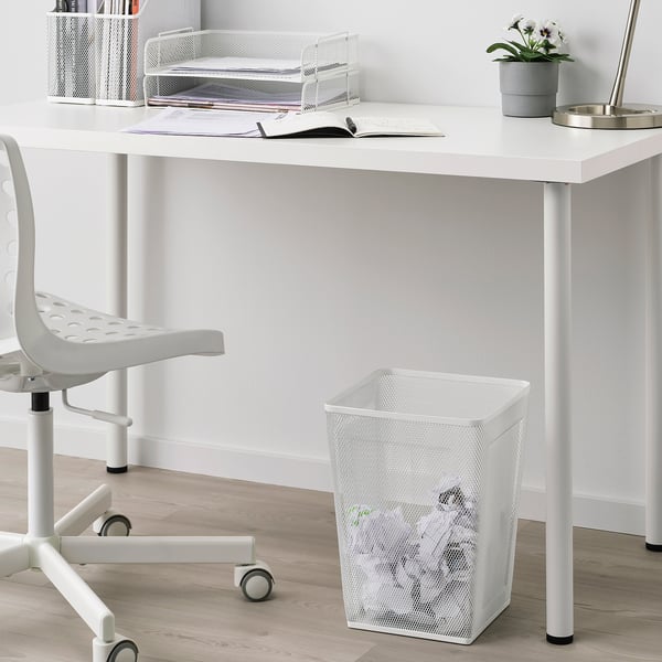 White metal mesh wastepaper basket under desk next to chair. Simple modern design. Smaller than chair and desk.