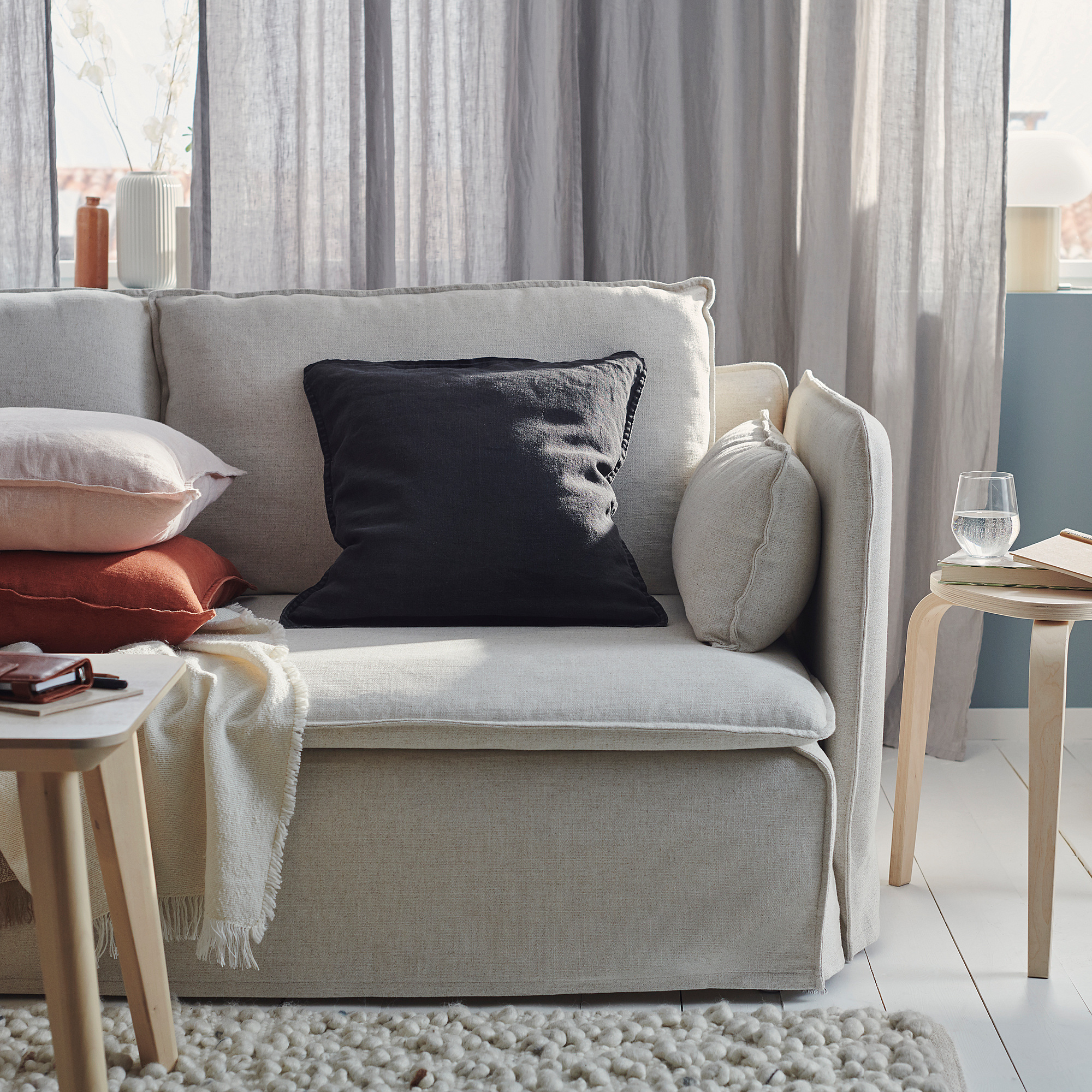 A cozy gray couch with black, gray, and peach pillows, a cream throw, wooden side tables, and a glass of water. Light gray curtains in the background.
