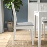 White wooden chair with grey cushion on patterned rug near potted plant.