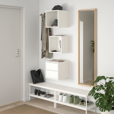 White wall-mounted EKET storage units display and hide items. Size and shape vary, organized next to a full-length mirror and plant.