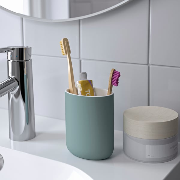 A green toothbrush holder on a white sink, containing two toothbrushes and toothpaste, is next to a metallic soap dispenser and a chrome faucet.