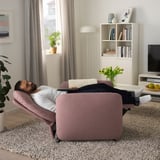 Man reclines in pink EKOLSUND; adjustable couch in cozy living space, next to a bookshelf and tv.