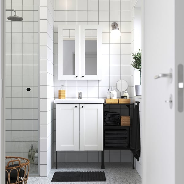 Modern minimalist bathroom with white ENHET vanity, mirror cabinet, and black towel rack. Glass shower, wooden stool, and grey tiled floor.
