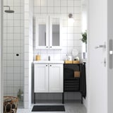 Modern minimalist bathroom with white ENHET vanity, mirror cabinet, and black towel rack. Glass shower, wooden stool, and grey tiled floor.