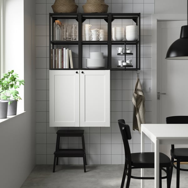 Modern kitchen with ENHET white cabinets, black open shelves, stacked dishes, and books. A black chair and stool are nearby.