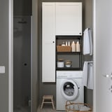 Modern bathroom with white ENHET cabinets, washing machine, open shelves with laundry supplies, towels, and a black shower in the background.