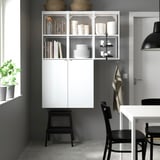 Minimalist kitchen: white ENHET shelving unit, with stacked dishes & glasses, books, & hanging towels; black chair & stool nearby.