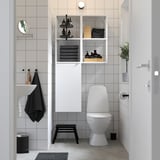 Modern bathroom with ENHET shelves, white/to gray tiles, toilet, sink, black stool, mirror, plant.