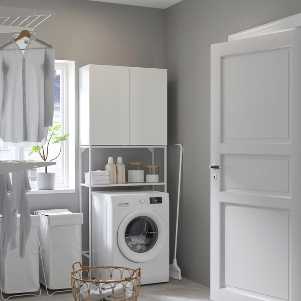 Laundry room with ENHET white cabinet storing washing machine, cleaners, and towels. By window, clothes hang on rack. Door closed.