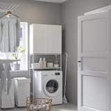 Laundry room with ENHET white cabinet storing washing machine, cleaners, and towels. By window, clothes hang on rack. Door closed.