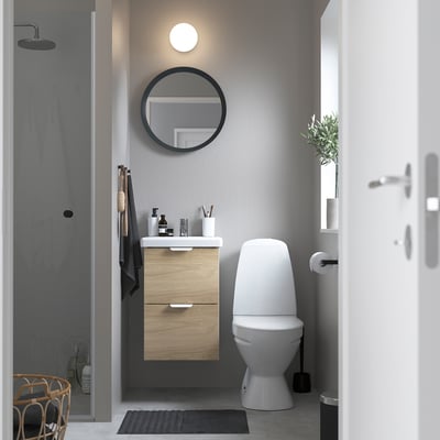 Modern minimalist bathroom with wall-mounted oak-like vanity, white toilet, round mirror, and grey wall.