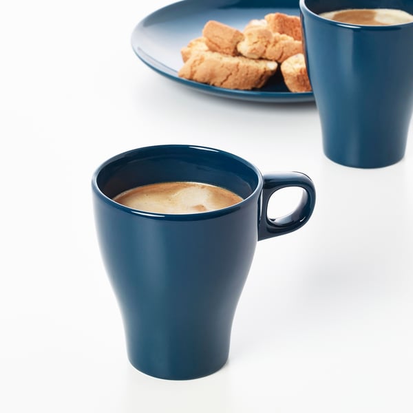 Two blue mugs filled with coffee, set on a white table, accompanied by a plate with biscuits.