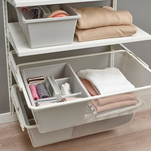 White organizer drawer with mesh basket holding small items in compartments. Folded clothes and accessories visible on shelves above.