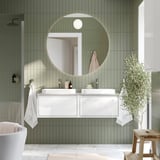 Modern bathroom with FAXÄLVEN round backlit mirror. Soft green tiles, white vanity, and plant decor. Mirror size notable, larger than turtle stool.