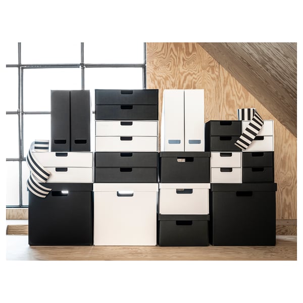 Stacked black and white storage boxes. Boxes vary in size, arranged neatly on wooden flooring and plywood wall. Modern and minimalist design.