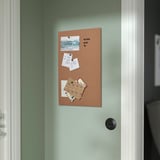 A cork memo board on a light green wall holds photos, notes, and cards with black pushpins, displayed vertically near a door.