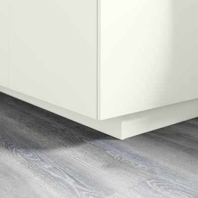 White kitchen cabinet plinth on gray wooden floor. Covers gap between floor and base cabinets. Part of FÖRBÄTTRA series.