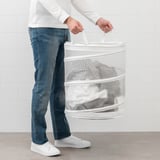 Person carrying white FYLLEN laundry basket, filled with folded clothes. Basket has handles for easy transport, allowing clothes to breathe.