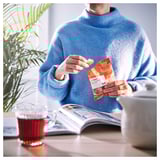 Person in blue sweater holding FYRTAKT candy, sitting at table with tea, plant, and magazine.
