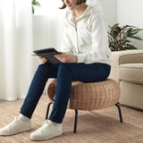 Woman sits on GAMLEHULT rattan footstool, holding a tablet, in a cozy room with natural light and plants.