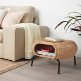 Beige GAMLEHULT rattan footstool with books & red box, next to sofa with pillows & plant. Compact & functional.