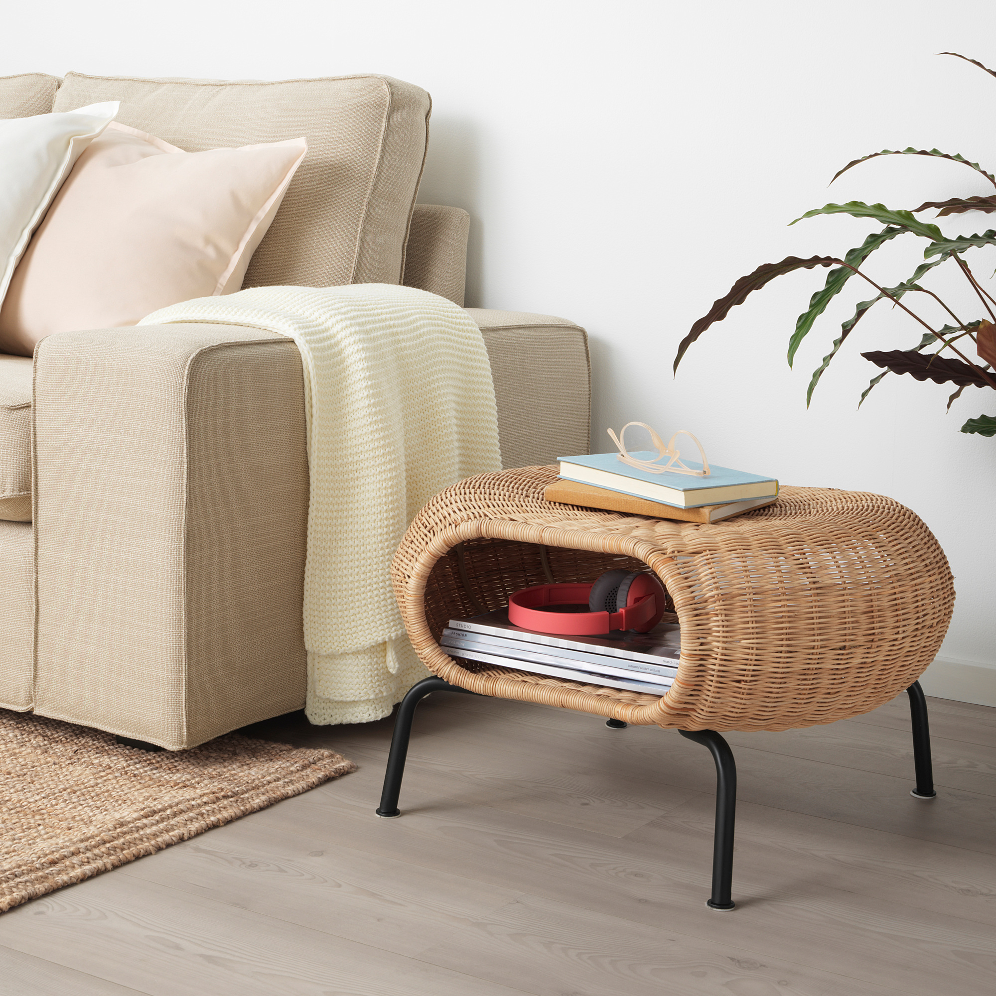 Beige GAMLEHULT rattan footstool with books & red box, next to sofa with pillows & plant. Compact & functional.