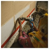 A woman is sewing a beige and red striped rug by hand. She uses a blue tool and wears colorful clothing.