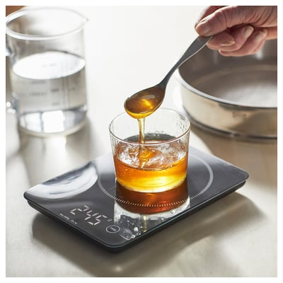 A digital kitchen scale, black with a glass top, measuring a liquid in a glass cup.