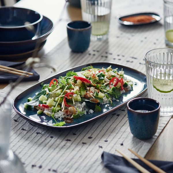 Blue glazed plate with rice and veggies on table with cups and chopsticks.