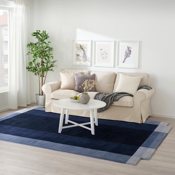 Living room with GNETGRÄS rug, blue shades, wool. Sofa, coffee table, and plant beside. Rug large, fits under furniture.