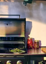 Black gas grill with silver trim and side burner. Vegetables cook in a pan on the grill. Red jars are nearby.