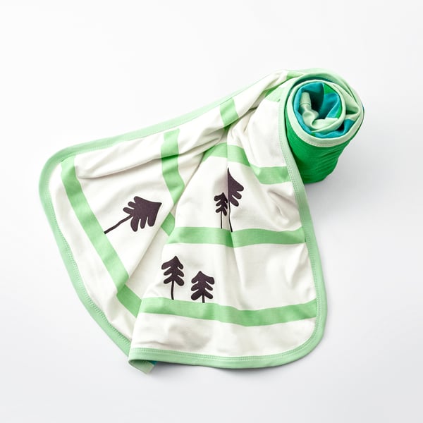 Baby blanket with soft, rolled edge, featuring forest trees on one side, laid out on a light grey surface.